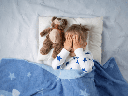 A little boy laying down to go to sleep with a teddy bear. He has hands over his eyes.