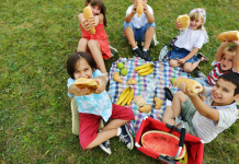 Six kids sitting around a tablecloth on the grass. They are happy and holding up the sandwiches they are eating.