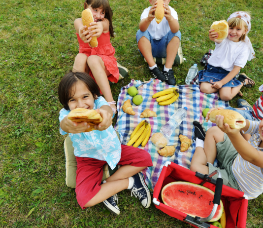 Six kids sitting around a tablecloth on the grass. They are happy and holding up the sandwiches they are eating.
