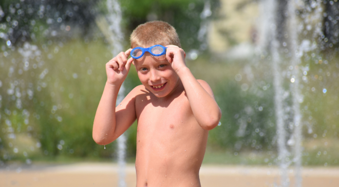 A young boy with blue swim goggles at a splash pad on a sunny day.