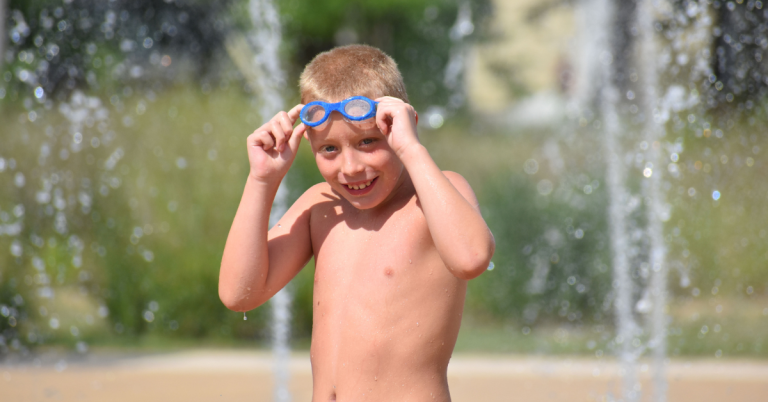 A young boy with blue swim goggles at a splash pad on a sunny day.