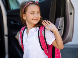 A young girl smiling and waving as she gets into a car. She is wearing a white shirt and has a bright pink back pack.