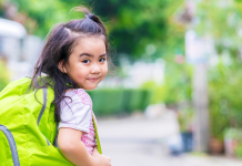 A little girl with a green backpack looking backwards over her shoulder and grinning.