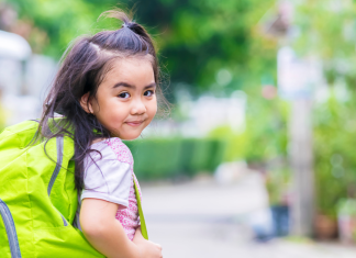A little girl with a green backpack looking backwards over her shoulder and grinning.
