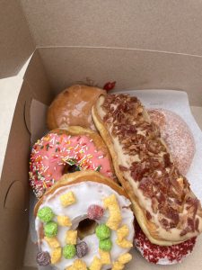 a box filled with half a dozen donuts of various flavors with toppings such as rainbow sprinkles, captain crunch cereal, and crumbled bacon