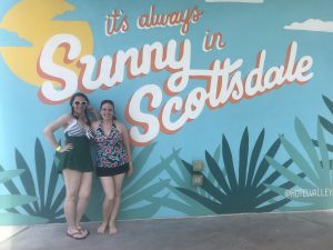 two women in swimsuits stand-in front of a mural that looks like a postcard that says it's always sunny in scottsdale
